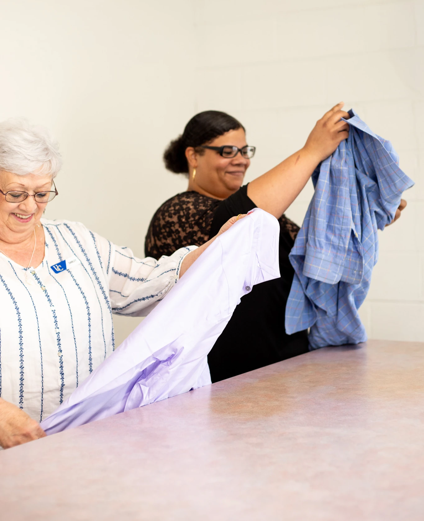 Two women stand at a table folding shirts, with one holding a light purple shirt and the other holding a blue plaid shirt.