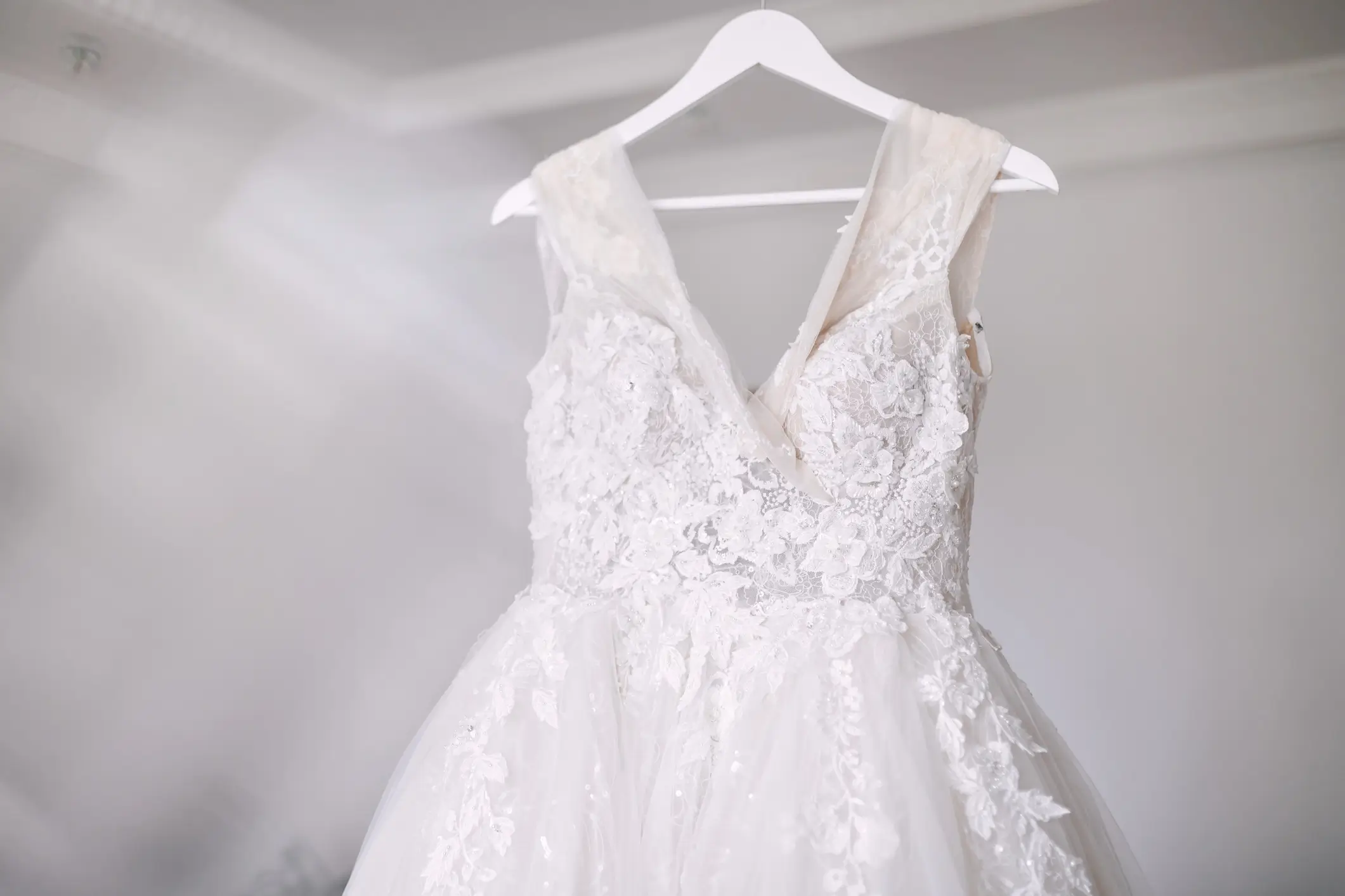 A white lace wedding dress with a tulle skirt hangs on a white hanger against a light, blurred background.